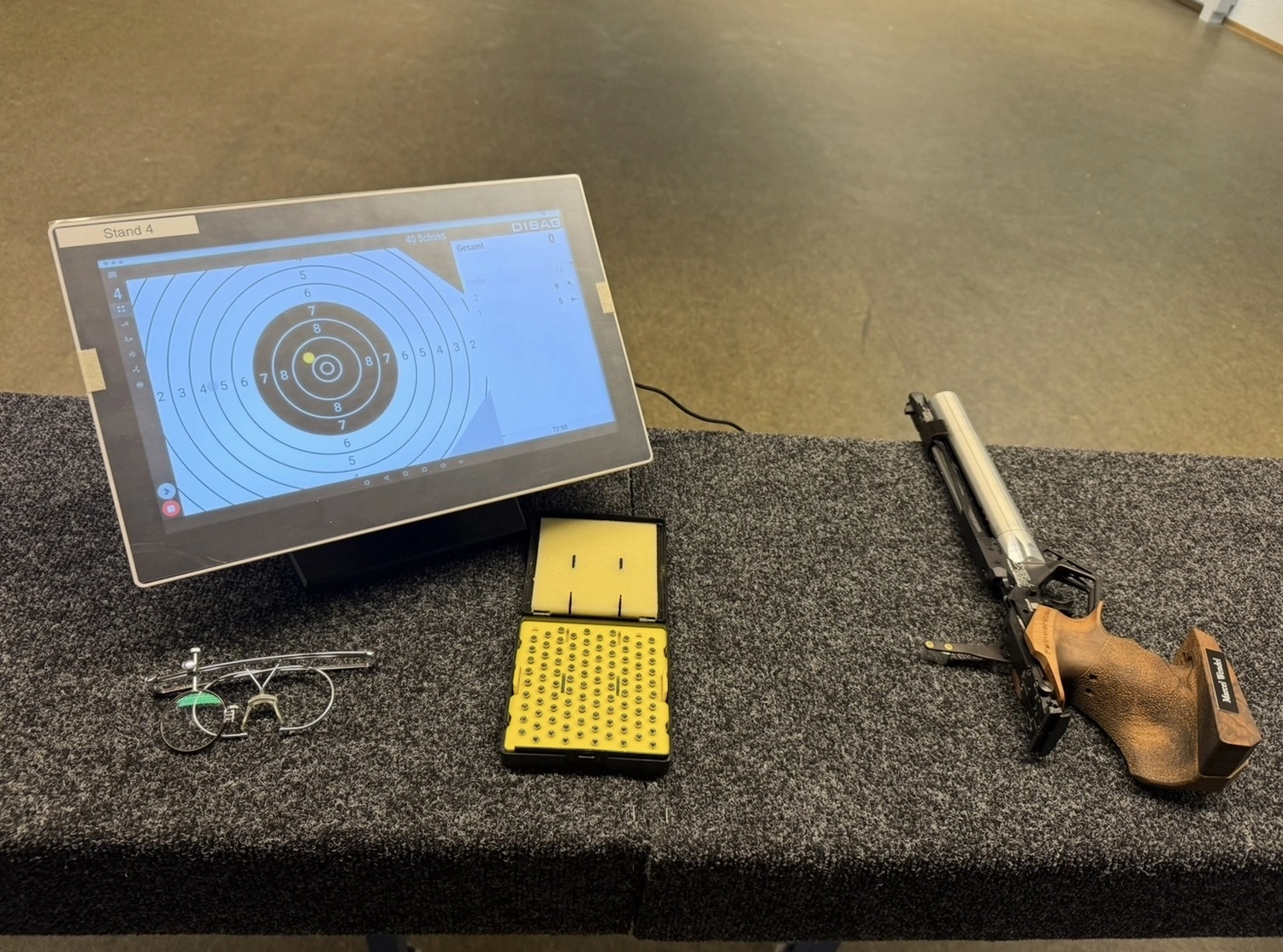 Schützenverein Bissingen/Enz e.V. – Schießausrüstung auf einer Bank: eine Sportpistole, eine Schachtel mit Schrotkugeln, eine Schießbrille und ein digitaler Monitor, auf dem eine Zielscheibe angezeigt wird.