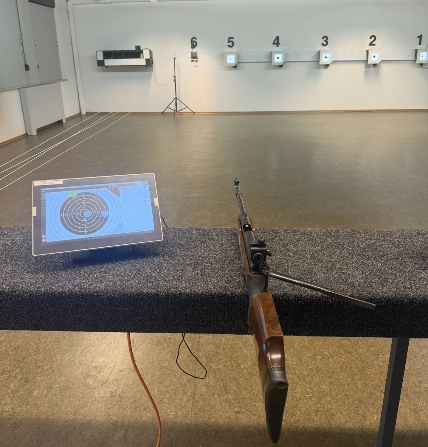 Schützenverein Bissingen/Enz e.V. – Eine Indoor-Schießanlage mit einem Gewehr und einem Tablet, auf dem eine Zielscheibe auf einer Bank zu sehen ist, sowie nummerierten Bahnen im Hintergrund.
