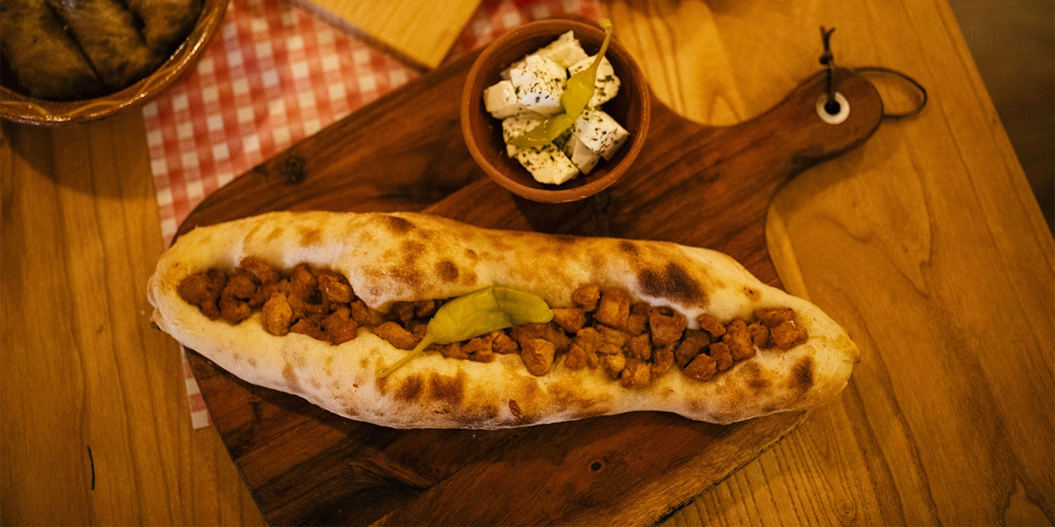 Schützenverein Bissingen/Enz e.V. – Ein bootförmiges Fladenbrot, belegt mit Fleischwürfeln und einer Paprikaschote, serviert mit einer kleinen Schälchen Feta-Käse und Paprikastreifen auf einem Holzbrett.